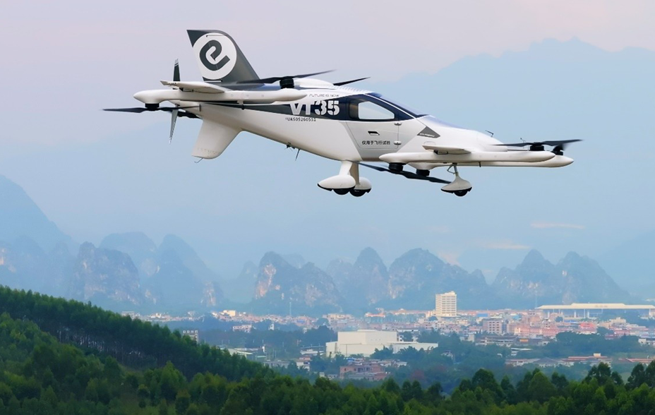 A sleek, white aircraft with multiple propellers flies over the lush, green landscape with distant mountains and a city skyline under a clear, pale blue sky.