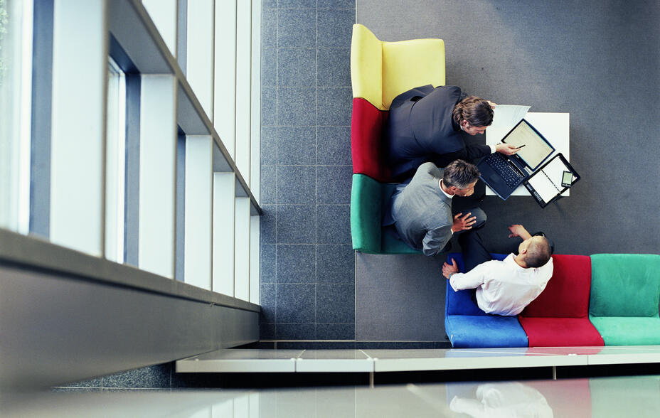 Three colleagues huddle over laptops, discussing strategy in a modern office lounge.