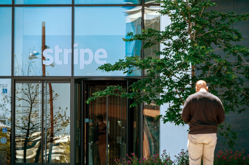 A person standing in front of a tree, outside a glass office block with the Stripe name printed on the glass