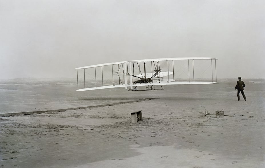 a monochrome image of the Wright brothers’ Flyer making the maiden flight above the sand dunes of Kitty Hawk, North Carolina
