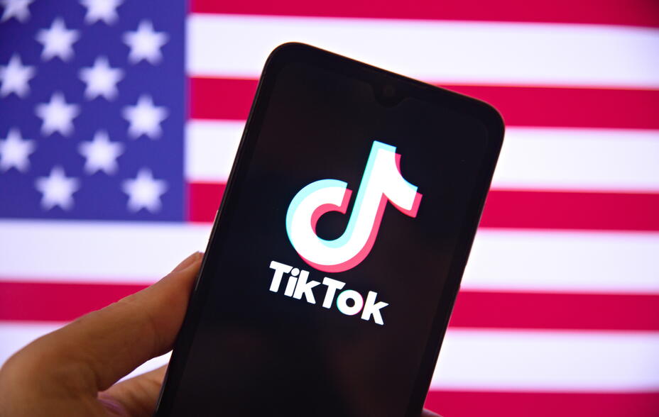 Hand holding a smartphone with the TIKTOK application logo on the screen and the United States flag in the background.