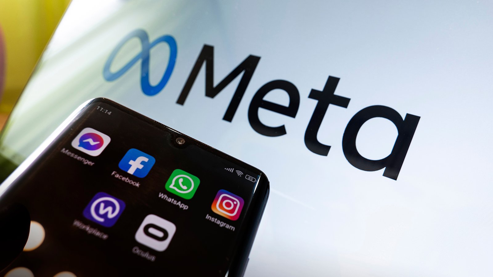 A smartphone, with the icons of the companies belonging to Facebook Metaverse, on the screen, held in front of a backdrop of the Metaverse logo.