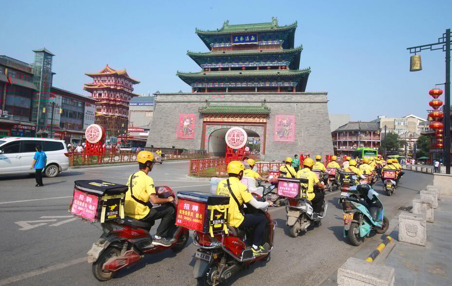 A group of food delivery couriers ride electric bikes, while wearing yellow vests, to deliver meals for Meituan Dianping in the ancient Kaifeng city.