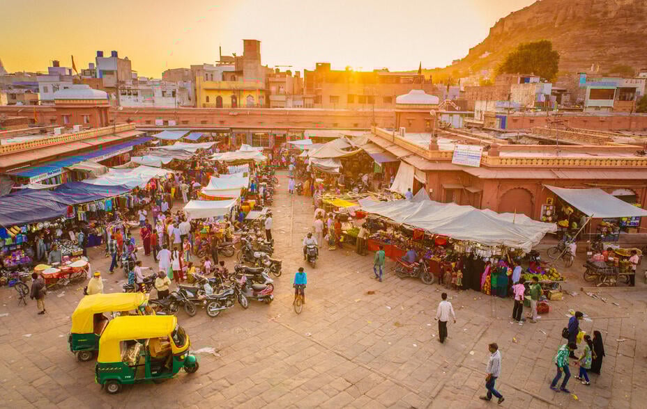 A bustling market in Jodhpur’s Old City at sunset. People walk past brightly coloured stalls and yellow rickshaws parked in the foreground.