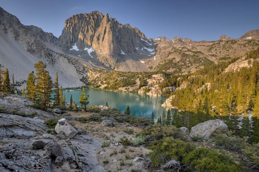 A calm turquoise lake sits in the valley between jagged mountains, the landscape peppered with evergreen trees.