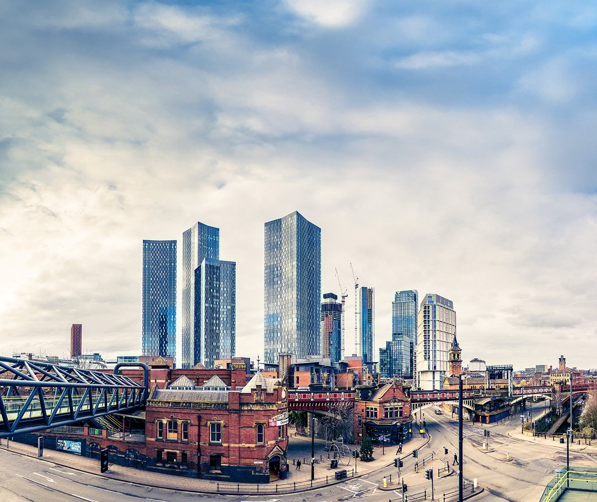 Panoramic view of Manchester city skyline in Lancashire, featuring tall glass skyscrapers under a cloudy sky, with historic red-brick buildings and a pedestrian bridge in the foreground.