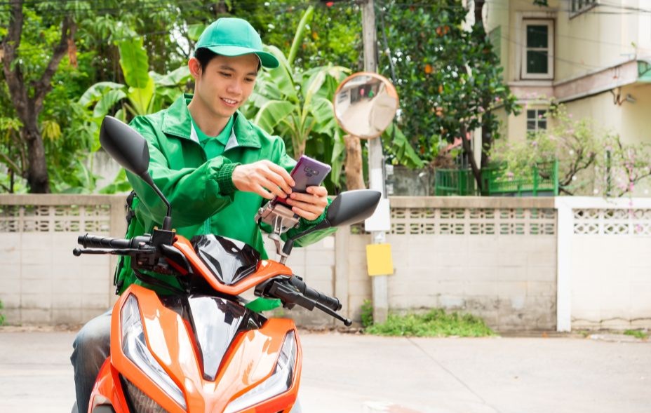 An Asian delivery man using an app on his mobile phone, straddling a motorcycle and wearing a green uniform.