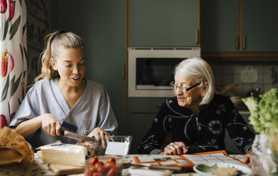 Female caregiver cutting bread loaf at dining table while sitting by senior woman in kitchen