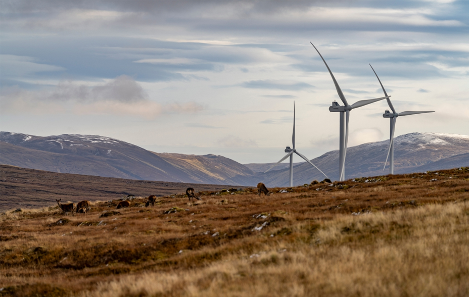 A mountainous Scottish scene; white wind turbines turn beside Highland deer.