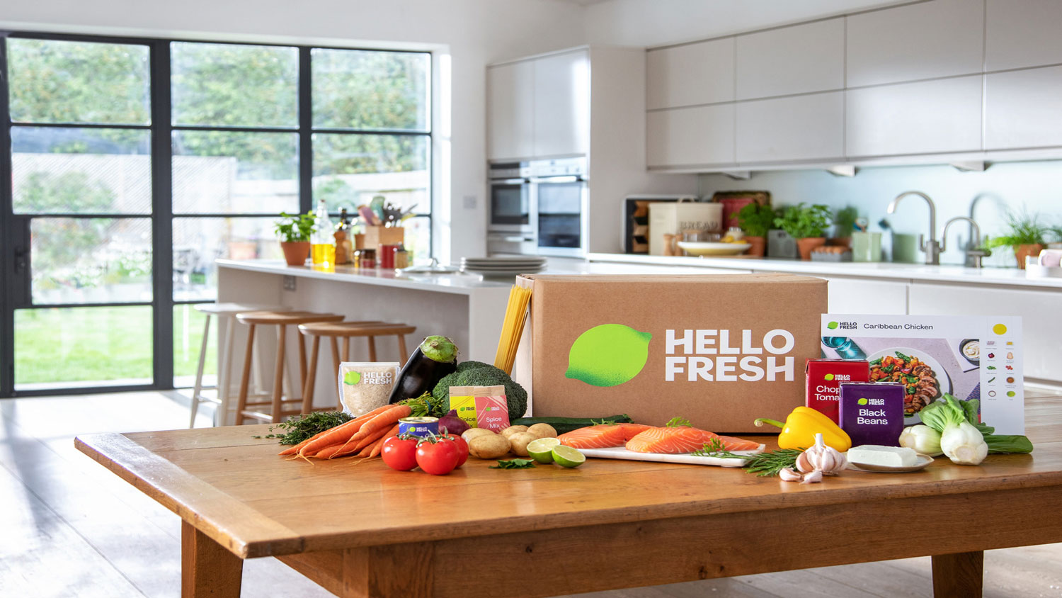 A Hello Fresh branded box surrounded by fresh produce on a table inside a kitchen.