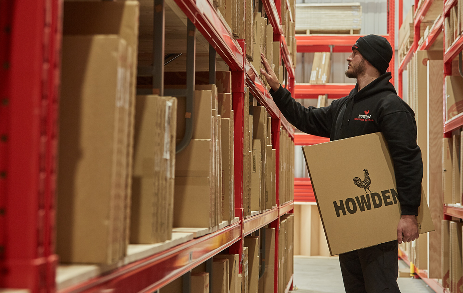 A man stands in a warehouse holding a flattened cardboard box under his arm. The Howdens logo is visible on the cardboard.